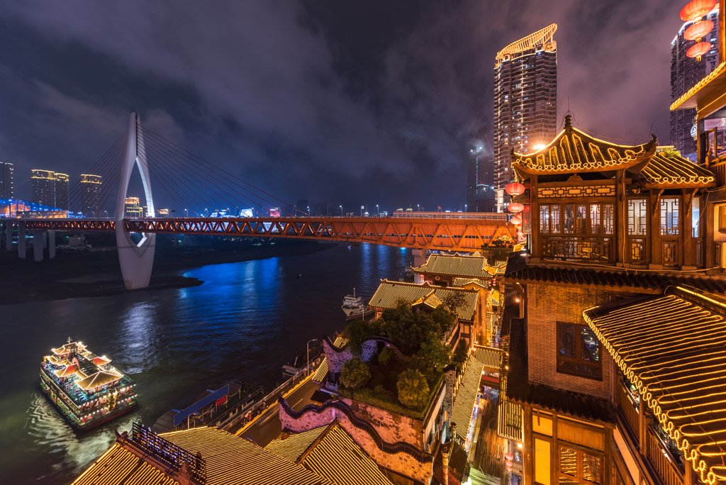 Hongya cave high angle view at night with Qiansimen bridge and an illuminated boat in the river, Chongqing, China