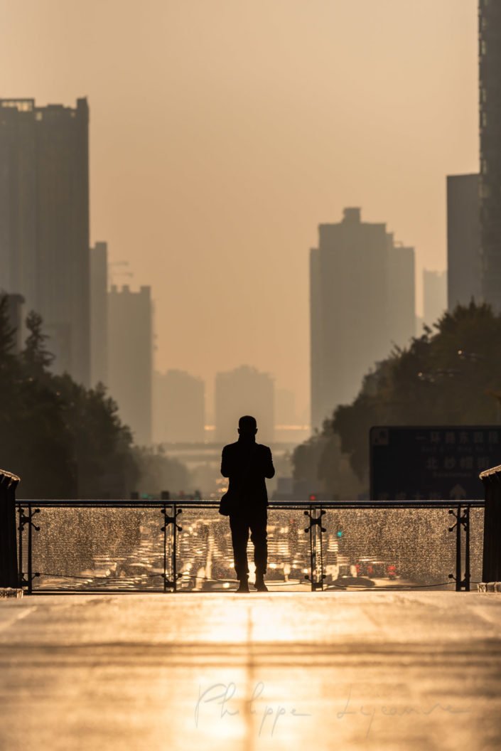 Man standing on a foot bridge at sunrise in downtown Chengdu city with car traffic in the background, Sichuan province, China