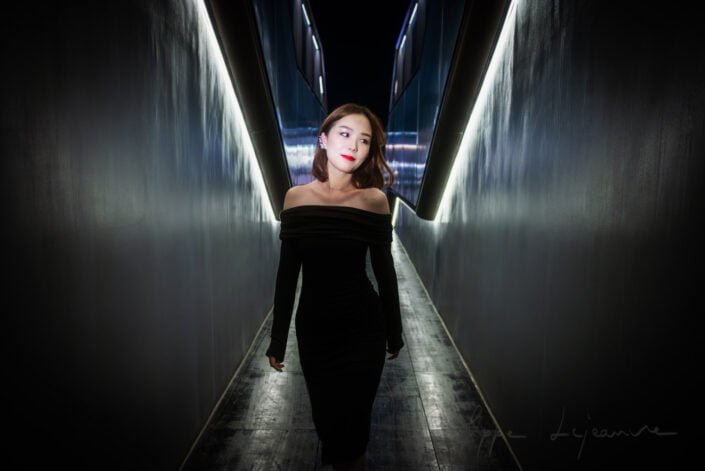 This image captures a moment of elegance and motion within the modern architectural space of SKP Chengdu, Sichuan province, China. A woman dressed in a black off-shoulder dress stands out against the dark, reflective background, her red lipstick adding a vibrant contrast. SKP, Chengdu, Sichuan province, China