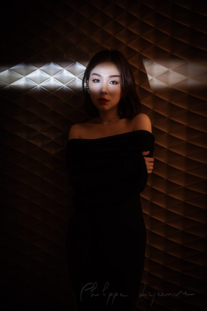 The image depicts a woman in a sophisticated pose, wearing an elegant black off-shoulder dress, against a textured wall with geometric patterns. Her makeup is polished, and she has a poised expression. SKP, Chengdu, Sichuan province, China