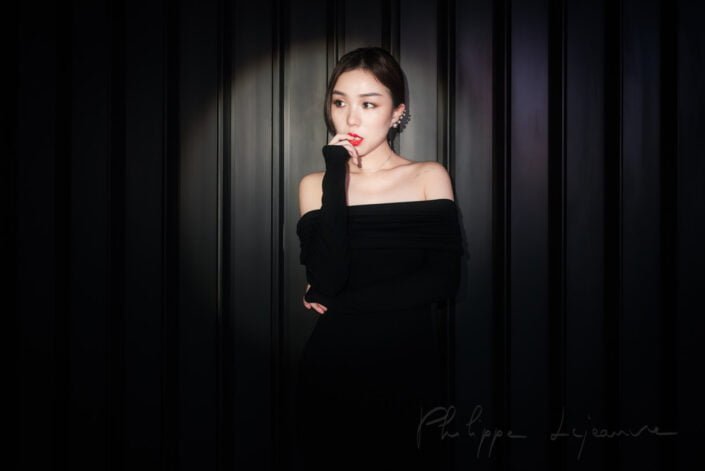 In this image, we see a woman poised in a moment of quiet reflection. Dressed in a chic black off-shoulder dress, she stands against a backdrop of vertical lines that add depth to the composition. The play of light and shadow accentuates her features, drawing attention to her thoughtful expression. Model : Fanny, Location: SKP, Chengdu, Sichuan province, China