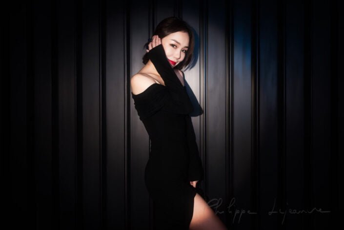 This image captures a woman exuding elegance and sophistication against a dark, vertical-striped background. Her attire is a classic black dress with an off-the-shoulder neckline, complemented by her red lipstick, which adds a pop of color to the otherwise monochromatic setting. Model : Fanny, Location: SKP, Chengdu, Sichuan province, China