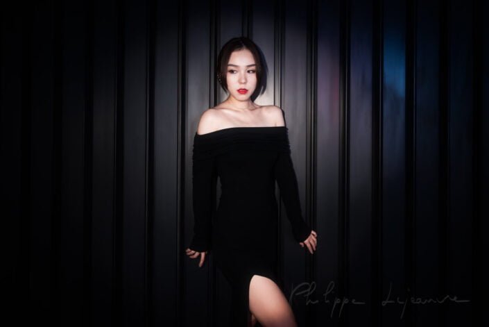 This image captures a woman exuding elegance and sophistication against a dark, vertical-striped background. Her attire is a classic black dress with an off-the-shoulder neckline, complemented by her red lipstick, which adds a pop of color to the otherwise monochromatic setting. Model : Fanny, Location: SKP, Chengdu, Sichuan province, China