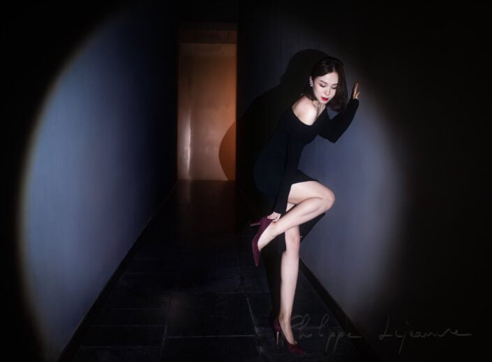 The image showcases a woman posed elegantly in a dimly lit hallway, characterized by her black attire and striking red lipstick, which stands out against the subdued background. SKP, Chengdu, Sichuan province, China