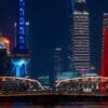 The photo features a striking nighttime panorama of Shanghai's Lujiazui area, highlighting the city's architectural prowess with towering skyscrapers adorned in colorful lights. Prominent structures such as the Oriental Pearl Tower and the Jin Mao Tower stand out against the dark sky, while the red Monument to the People's Heroes adds a splash of color to the urban landscape. The steel truss of Waibaidu bridge in the foreground provides a sense of scale and depth, leading the viewer's eye across the water towards the cityscape. The overall mood is one of awe-inspiring modernity and cultural significance, encapsulating the dynamic energy of China's financial hub.