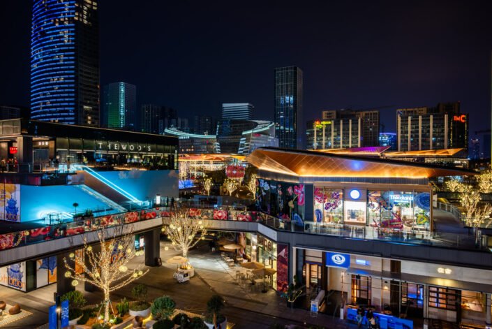 Nocturnal scene showcases the bustling commercial hub of Baoli ShiGuangLi in Chengdu, China. The skyline is adorned with various illuminated buildings, including a standout circular structure showcasing colorful graphics.