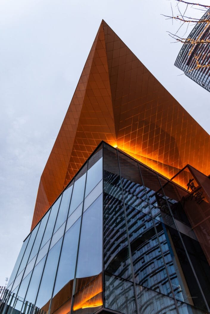 Modern building on the Baoli ShiGuangLi complex in Chengdu, Sichuan province, China. The building's design features a bold, angular composition with a prominent golden-orange triangle at its apex, creating a stark contrast with the reflective glass facade below. The low angle of the shot emphasizes the building's grandeur and the intriguing play of light on its surfaces. The sky is partly cloudy