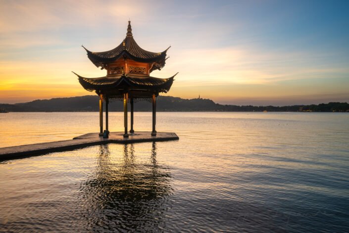 A harmonious blend of natural and man-made elements, highlighting the pavilion's intricate design with its golden illuminated roof and dark wooden pillars. The calm water reflects the pavilion's silhouette, creating a mirror-like effect that enhances the scene's tranquility. The sky transitions from warm orange hues near the horizon to cooler blues as it stretches upward, adding depth and contrast to the composition. The overall mood is one of peacefulness and contemplation, inviting viewers to appreciate the cultural significance and aesthetic elegance of this iconic landmark.in Hangzhou, Zhejiang province, China