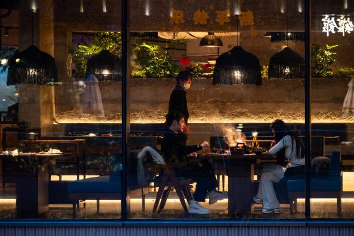 Intimate ambiance of a restaurant, the image reveals customers comfortably seated inside. The interior is warmly lit, featuring black hanging lanterns that cast a soft glow and enhance the tranquil dining experience. The exterior display screen, adorned with Chinese characters, adds a touch of authenticity. Outside, passersby glance at the inviting scene, encapsulating the lively street culture of Chengdu. The window view elegantly balances between the indoor warmth and the outdoor curiosity. The establishment's name suggests it may specialize in traditional Chinese cuisine. Illuminated signs and modern interior elements contrast against the subdued street lighting, creating an appealing visual narrative of urban life and gastronomy.