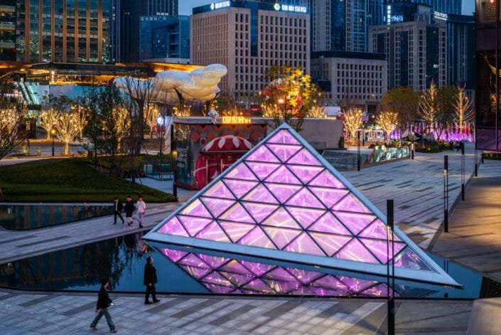 Modern illuminated geometric structure amidst a tranquil urban park in the Baoli ShiGuangLi area of Chengdu. The artificial lighting highlights the structure and reflects beautifully on the surrounding surroundings. Several people are seen walking around