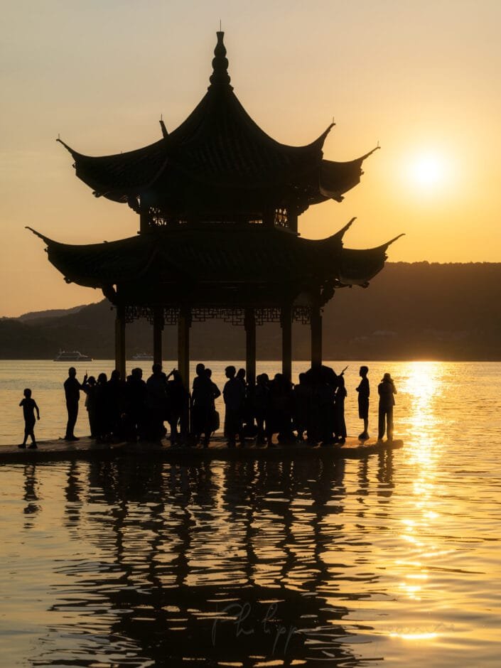 A breathtaking moment as the sun sets over Xihu - West lake - in Hangzhou, Zhejiang province, China. The silhouetted figures stand before the iconic Jixian Pavilion, its intricate rooflines and tiered eaves creating an elegant reflection on the shimmering water surface. The warm hues of the setting sun cast long shadows and a soft glow that enhances the peaceful ambiance of this cultural landmark.