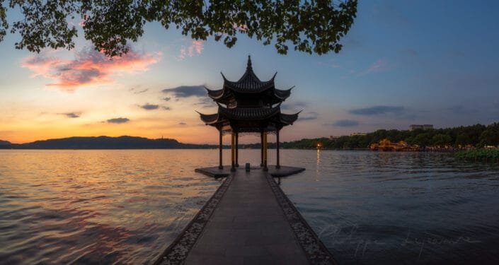 Timeless beauty of the Jixian Pavilion, with its intricate roof and elegant design, standing gracefully on a wooden pier that extends into the calm waters of the lake. The warm hues of the setting sun paint the sky in shades of pink, orange, and blue, creating a peaceful atmosphere. In the distance, silhouetted mountains add depth to the landscape, while the gently rippling water reflects the soft light of dusk. This serene tableau highlights the harmonious blend of natural and architectural elements, evoking a sense of tranquility and cultural heritage. Hangzhou, Zhejiang province, China