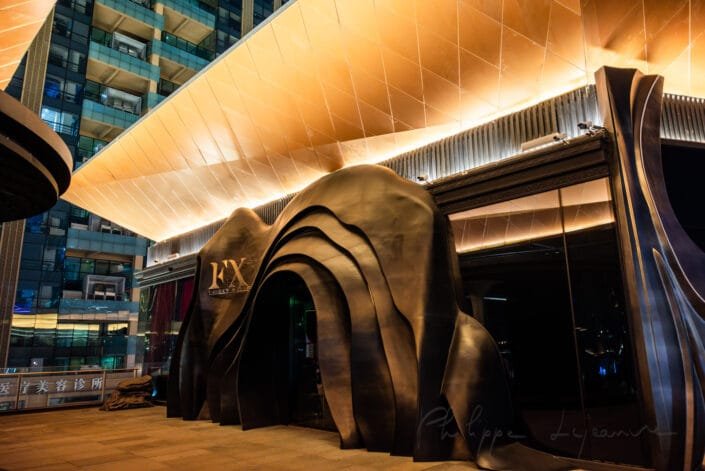 Modern, visually striking entrance to Baoli ShiGuangLi, a commercial complex in Chengdu, Sichuan province, China. The entrance features an artistic, flowing design that resembles natural rock formations, creating a sense of depth and movement. The structure is illuminated with warm, golden lighting, enhancing its elegance and sophistication. The surrounding architecture includes tall, glass-paneled buildings, reflecting the contemporary urban style of the area. The overall composition is balanced, with the entrance taking center stage, while the surrounding buildings provide a dynamic backdrop.