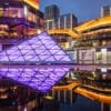 Urban nightscape at Baoli ShiGuangLi in Chengdu, Sichuan province, China. The central focus is a violet, pyramid-shaped structure illuminated with vibrant lighting, reflected on a glass surface in the foreground that mirrors the city's lights and buildings. Contemporary buildings with gold accents surround the area, adding to the futuristic atmosphere. This urban setting is illuminated with citylights, creating a serene yet energetic ambiance