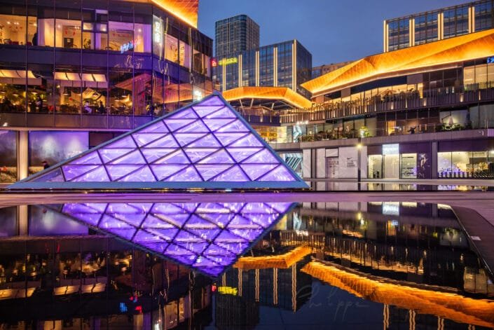 Urban nightscape at Baoli ShiGuangLi in Chengdu, Sichuan province, China. The central focus is a violet, pyramid-shaped structure illuminated with vibrant lighting, reflected on a glass surface in the foreground that mirrors the city's lights and buildings. Contemporary buildings with gold accents surround the area, adding to the futuristic atmosphere. This urban setting is illuminated with citylights, creating a serene yet energetic ambiance