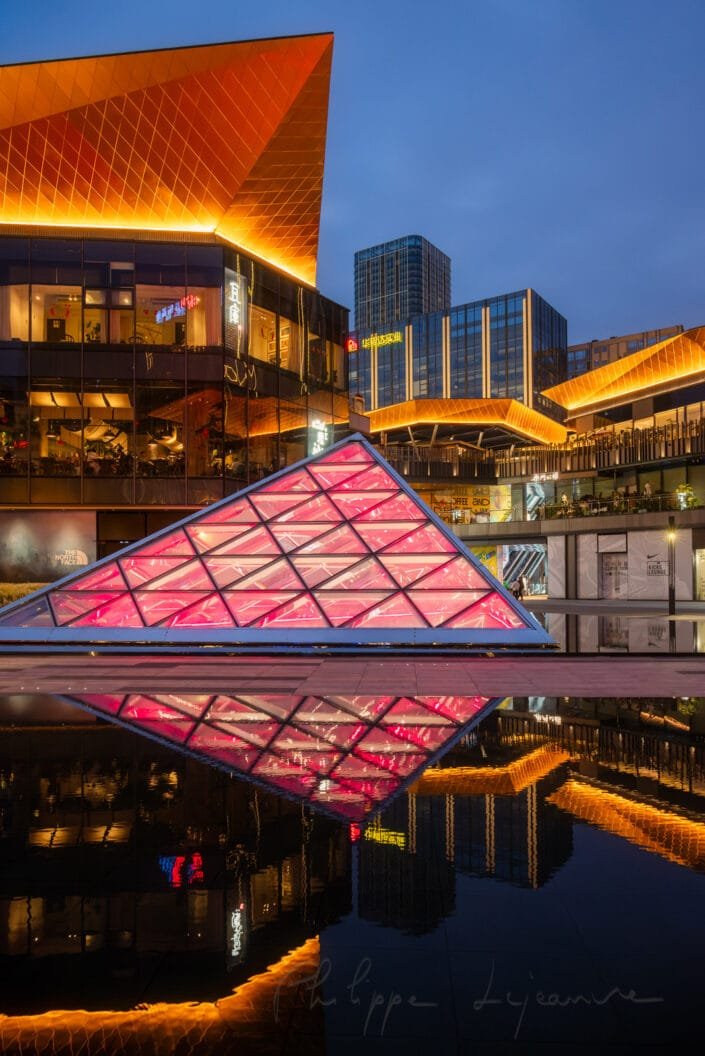 Urban nightscape at Baoli ShiGuangLi in Chengdu, Sichuan province, China. The central focus is a pink, pyramid-shaped structure illuminated with vibrant lighting, reflected on a glass surface in the foreground that mirrors the city's lights and buildings. Contemporary buildings with gold accents surround the area, adding to the futuristic atmosphere. This urban setting is illuminated with citylights, creating a serene yet energetic ambiance