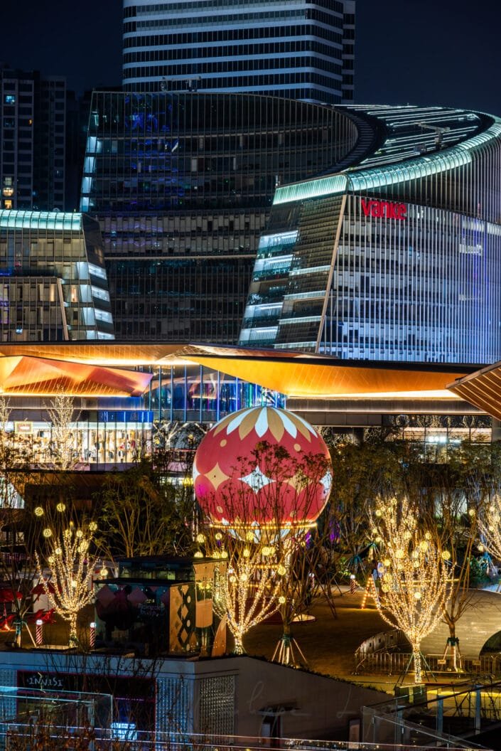 Bustling urban landscape of Chengdu's Baoli ShiGuangLi district comes alive at night, as traditional and contemporary aesthetics harmonize. A large red lantern with intricate white and gold patterns hangs prominently, surrounded by smaller white-lit trees, creating a magical ambiance. The photo showcases the city's fusion of cultural heritage and modern progress. Neon-lit skyscrapers and vibrant buildings serve as the backdrop to this picturesque scene