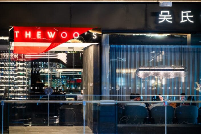 Exterior of a contemporary restaurant. The bold, red neon sign reading THE WOOS stands out prominently against the dark facade. Inside, patrons can be seen seated, suggesting a lively dining experience.