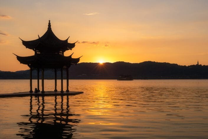 A silhouette of a beautifully designed pavilion stands proudly against a sky tinged with warm colors as the sun descends below horizon; its peaceful and scenic backdrop creates a moment of serenity.