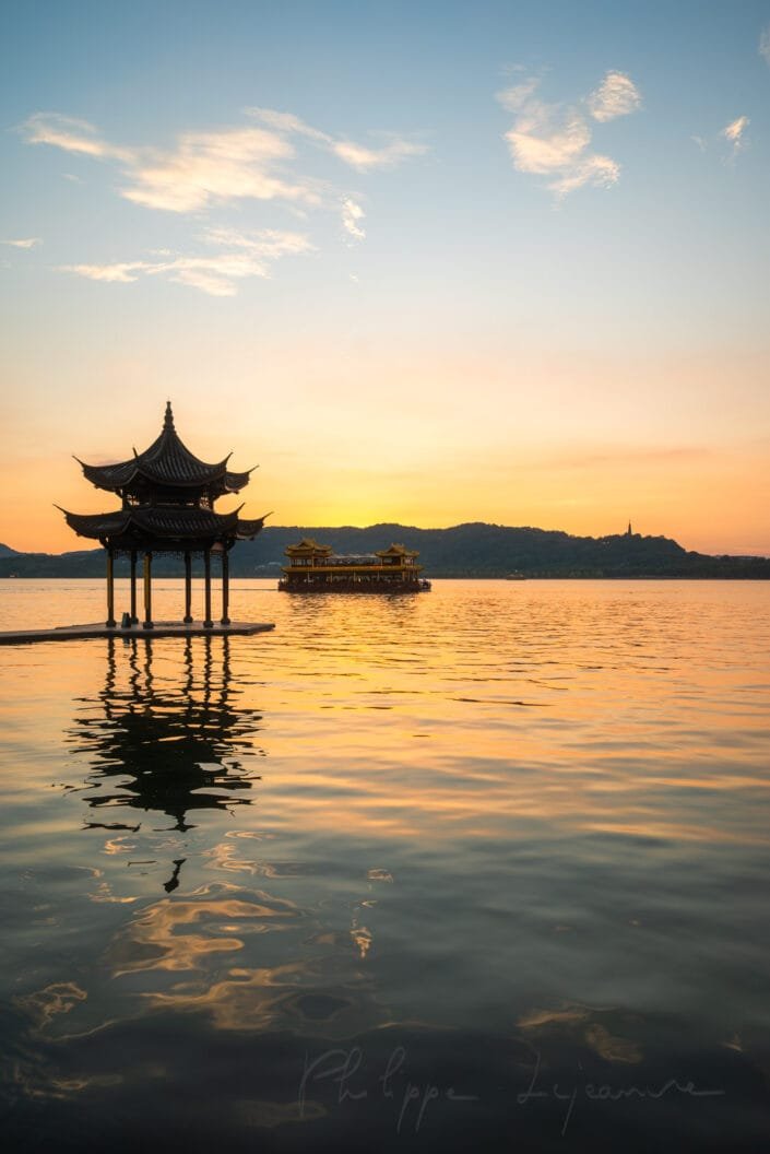 A silhouette of a beautifully designed pavilion stands proudly against a sky tinged with warm colors as the sun descends below horizon; its peaceful and scenic backdrop creates a moment of serenity.