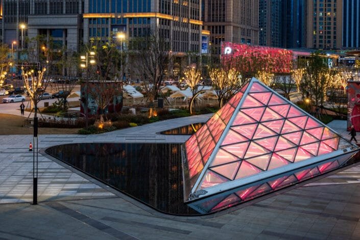 Modern pyramid glass structure featuring a diamond-shaped facade illuminated in pink at night, set under a cityscape with lit buildings and pedestrian activity in Chengdu