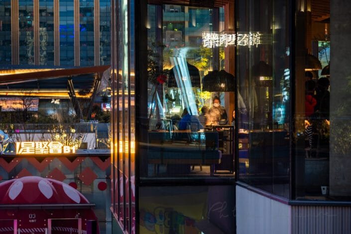 Vibrant nightlife at Baoli ShiGuangLi in Chengdu, Sichuan, showcasing patrons enjoying their meals in a brightly illuminated eatery. The scene is set against the backdrop of the city's architecture, with lights and silhouettes hinting at the bustling urban environment. The warm glow of neon signs and table lamps reflects off the glass facade, creating a welcoming ambiance. This snapshot of city life conveys a sense of cultural richness and the dynamic energy of urban dining.