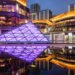 Urban nightscape at Baoli ShiGuangLi in Chengdu, Sichuan province, China. The central focus is a violet, pyramid-shaped structure illuminated with vibrant lighting, reflected on a glass surface in the foreground that mirrors the city's lights and buildings. Contemporary buildings with gold accents surround the area, adding to the futuristic atmosphere. This urban setting is illuminated with citylights, creating a serene yet energetic ambiance